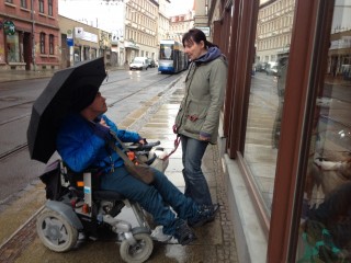 In leichter Sprache - Über einen Rundgang | nicht immer Rollstuhl gerecht. Daniela Nuß mit Besucher vor dem Laden.